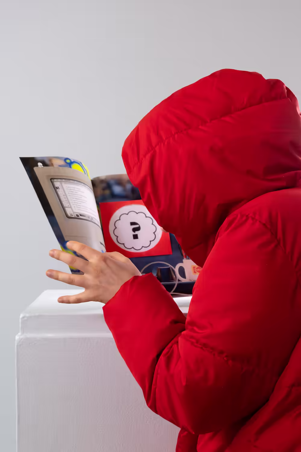 Figure in a red hooded jacket peeks into the Hijinx World magazine, revealing a secret message inside.