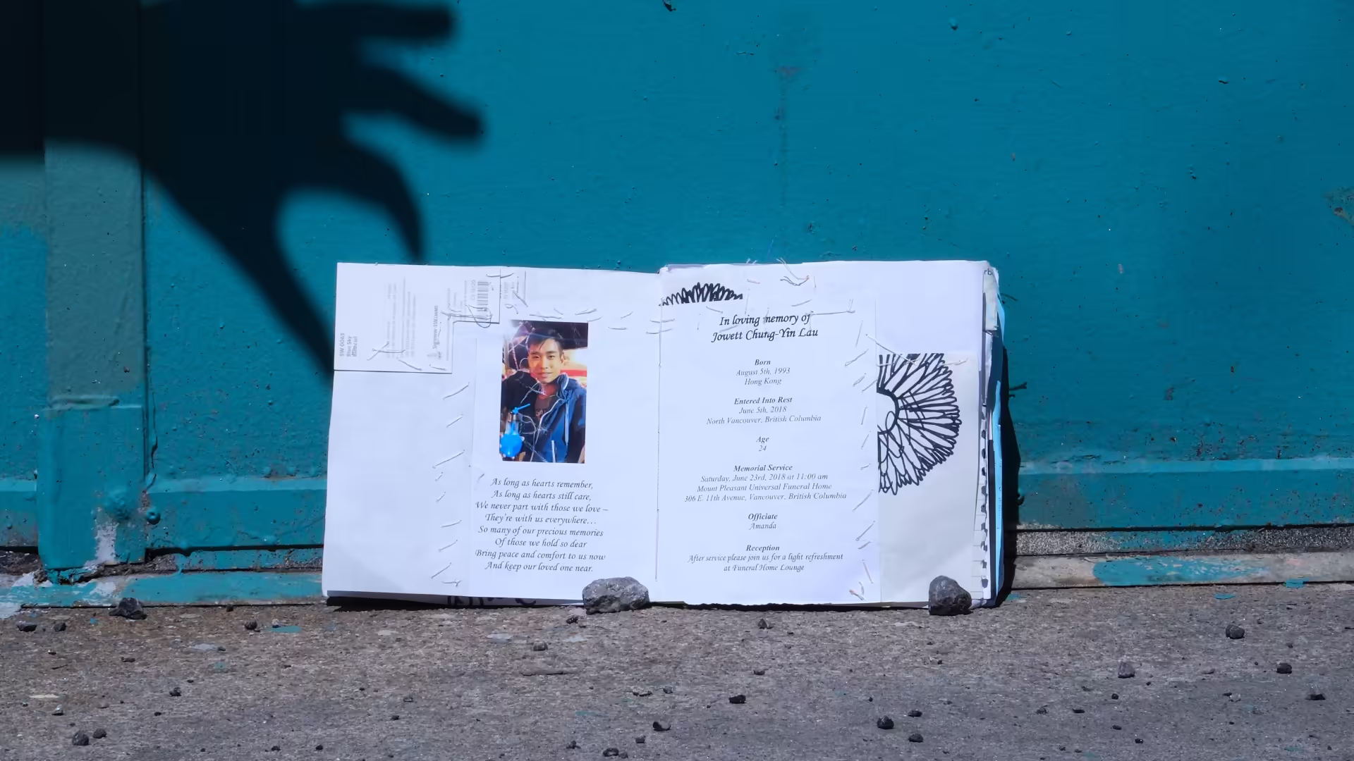 Journal sitting by a teal wall, with a shadow of a hand approaching. Open at page featuring an invite to a funeral.